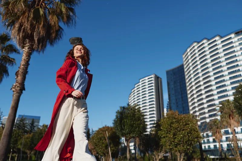 Young woman in a red coat walks confidently along a sunny urban street with modern buildings and palm trees in the background.