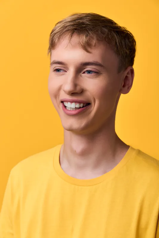 Smiling young man in a bright yellow shirt against a vivid yellow backdrop, friendly candid vibe, headshot style with studio lighting, clean background for lifestyle or fashion use
