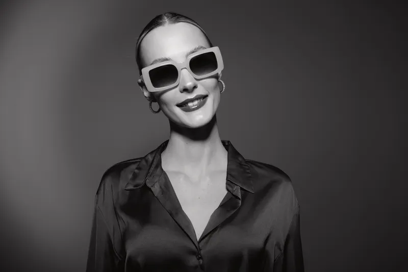 Young Caucasian woman with fair skin and sleek hair wears oversized sunglasses and a satin blouse, smiling confidently against plain studio background.