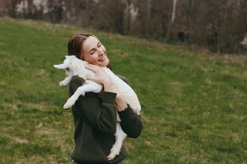 Joyful woman outdoors embraces a playful goat kid, warm sunlight, relaxed posture, gentle breeze, rustic pasture setting, candid moment, natural colors, wholesome lifestyle, connection with animals,