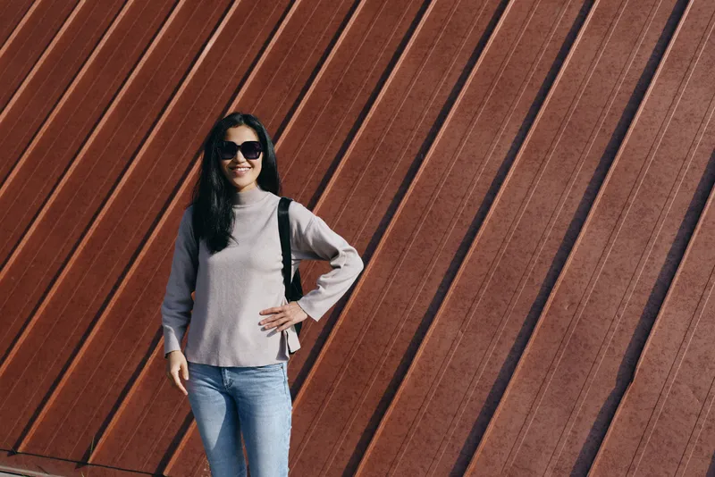 Woman wearing sunglasses and gray sweater poses against a diagonal wooden wall, outdoor street fashion with a confident stance and urban chic vibe, bright daylight