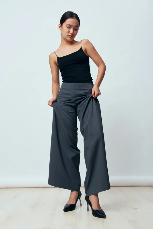 Young woman wearing black tank top and high waist wide leg gray pants standing indoors on light wooden floor with thoughtful pose and neutral light gray background.