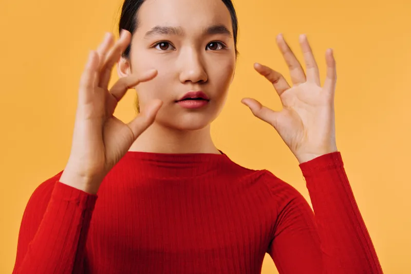Woman in red sweater posing with hands raised and facial expression on orange background. Asian female model showing fingers and engaging with camera indoors.