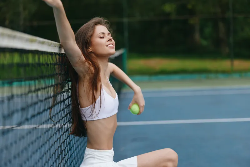 Young woman enjoying a tennis break, wearing a sports bra and shorts, smiling with tennis ball in hand against a vibrant green background, conveying athleticism and joy
