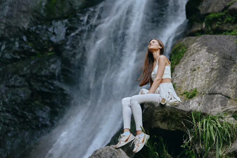 young woman sitting by waterfall enjoying nature, wearing white activewear and sneakers, serene outdoor scene with rocky background and flowing water