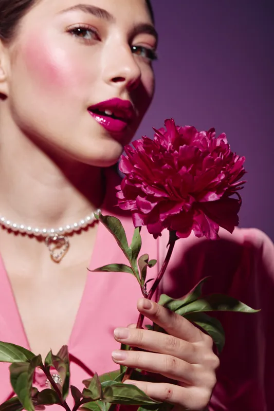 Young woman in pink outfit with pearl necklace holding magenta flower near her face. Studio shot with purple background. Concept of beauty creative fashion and elegance.