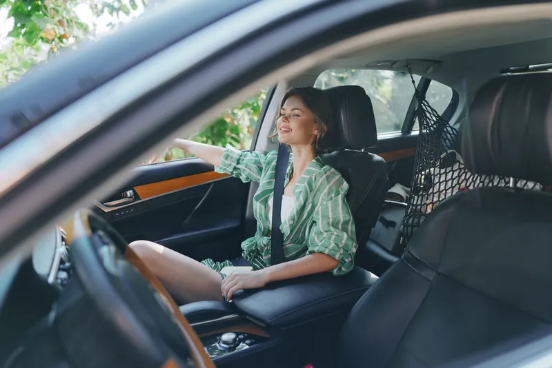 Portrait of a smiling woman driving a modern car, seated in the front seat with a relaxed pose and casual green striped shirt.