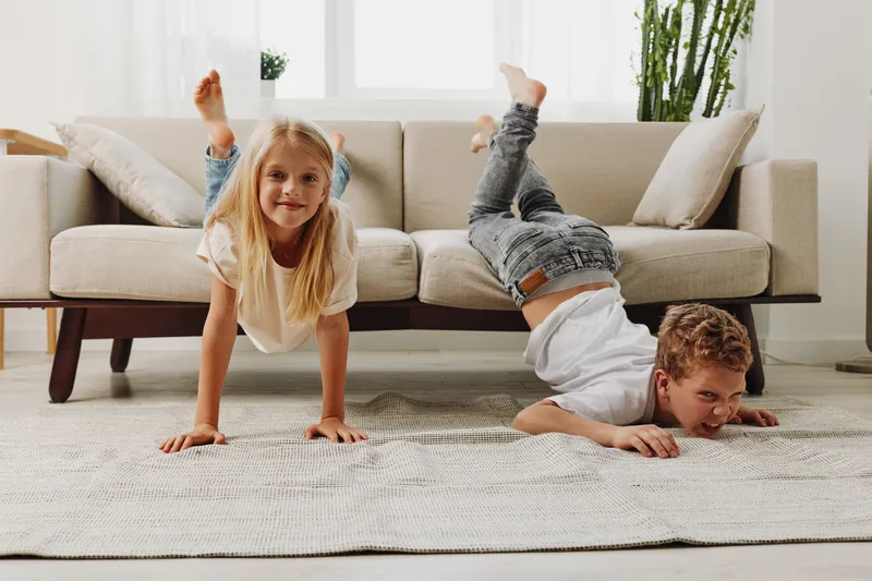 Two playful children exercise on a carpet in a bright living room with a sofa and plants, enjoying active indoor fun and sibling bonding.