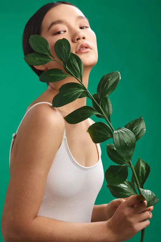 Asian woman poses with a leafy branch in a minimal studio, conveying calm confidence and natural beauty. Soft lighting and neutral attire create a clean, modern aesthetic for wellness branding.