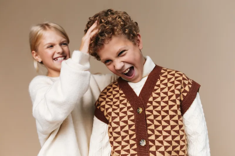 Two friends share a joyful moment in a cozy studio, one tugging the other's hair while both wear warm knitwear, bright smiles, and a soft beige backdrop.