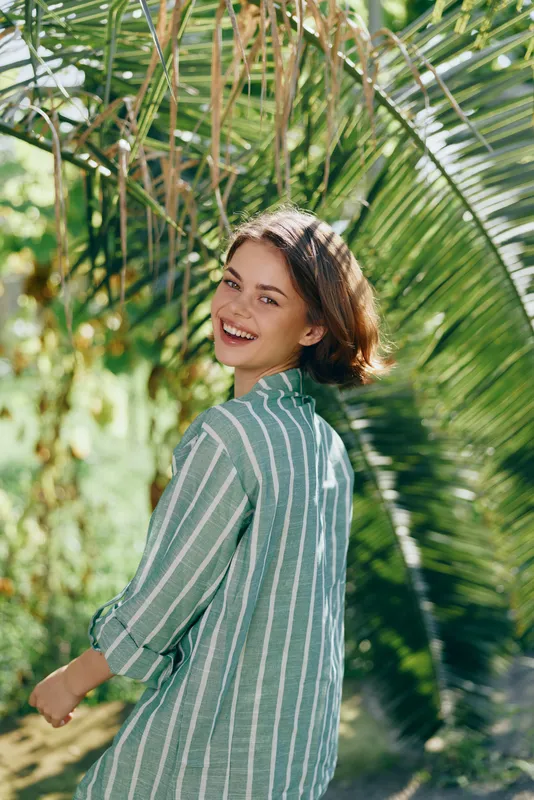 Description1: A smiling woman in a striped green shirt outdoors, bathed in sunlight among lush tropical greenery.