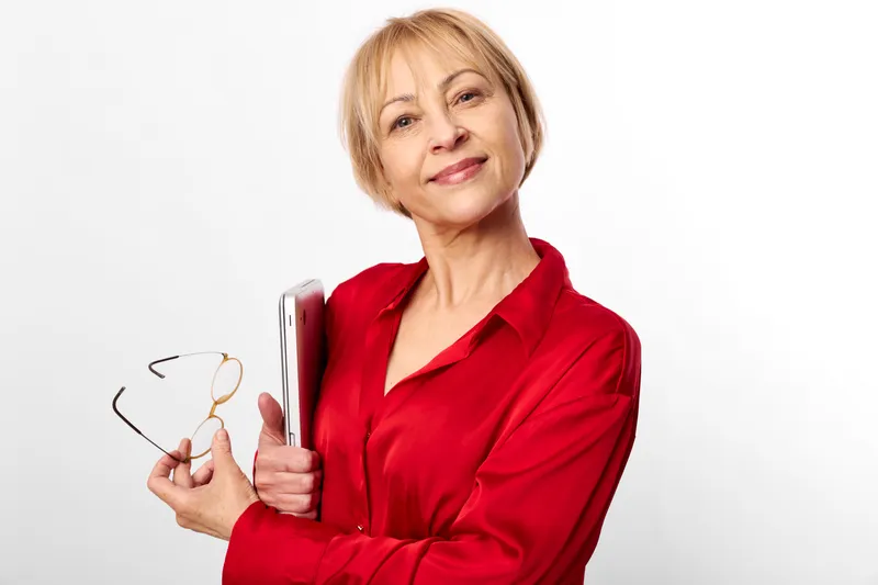 Senior woman holding eyeglasses and a book, smiling confidently in a red shirt on a white background. Reading lifestyle concept for vision and learning.