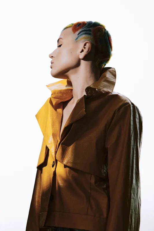 Profile of a woman with colorful short hair wearing a brown leather jacket, showing a stylish and modern look with a clean white background.