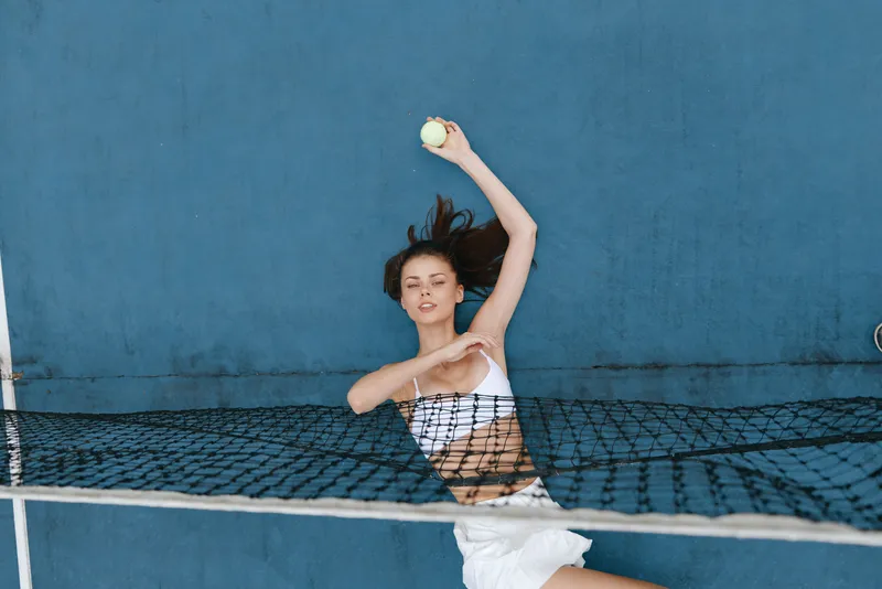 Young female tennis player in white sportswear serving a ball against a blue background, expressing confidence and energy, ideal for sports and wellness concepts