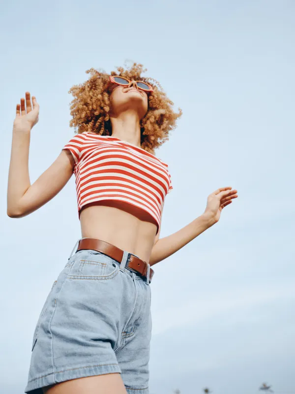 Young woman dancing outdoors in a red striped crop top and light denim shorts, smiling with carefree energy under a bright blue sky.