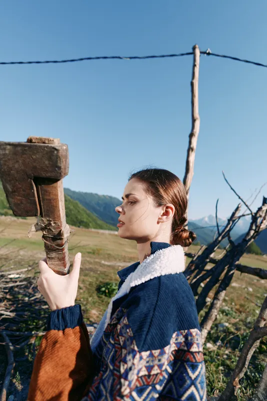 Woman portrait in countryside by a wooden fence, wearing patterned sweater in profile against mountains. Rural outdoors scene with sky, grass and serene seasonal mood.