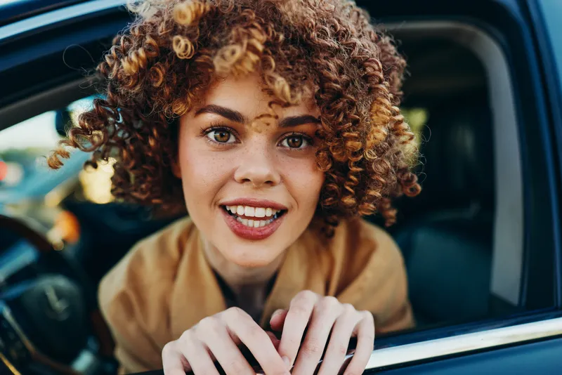 Joyful woman leaning from a car window, curly hair illuminated by warm sunlight, candid lifestyle moment, casual attire, bright colors, urban backdrop