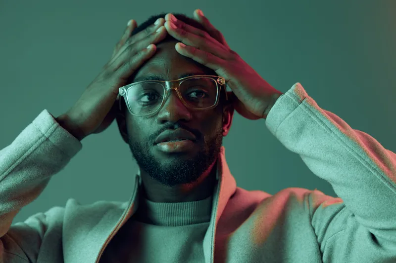 A thoughtful black man in stylish glasses and a light coat, expressing concern against a soft green and pink background symbolizes introspection and emotional depth