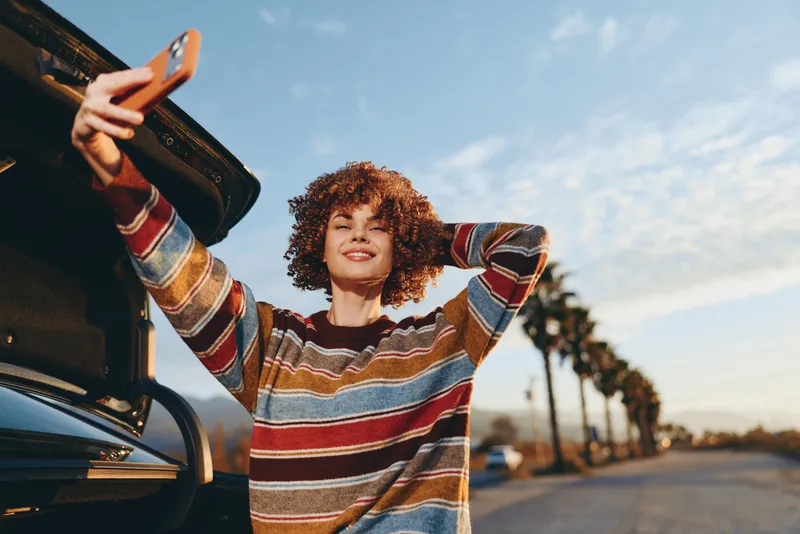 Woman lifestyle, curly woman holds phone near open car door on road at sunset, striped sweater, warm sunlight, relaxed smile, travel moment by vehicle