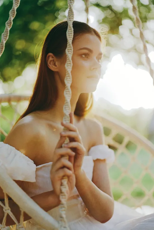 Serene young woman in a white off shoulder dress sitting on a swing, surrounded by nature, exuding tranquility and summer vibes, perfect for wellness and relaxation themes