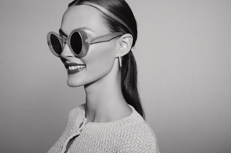 Smiling young Caucasian woman with fair skin and sleek hair wears large round sunglasses and a knitted sweater. Studio portrait with soft lighting and stylish fashion concept.
