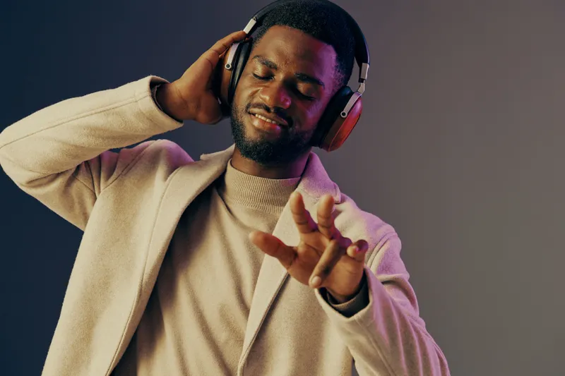A young Black man enjoying music with headphones in a stylish light colored jacket, expressing joy and relaxation in a modern, colorful background