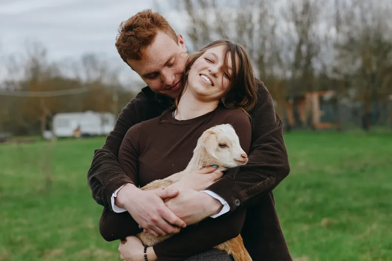Couple smiling as they embrace a goat kid in a meadow, a warm lifestyle moment blending affection, nature, and farm life outdoors.