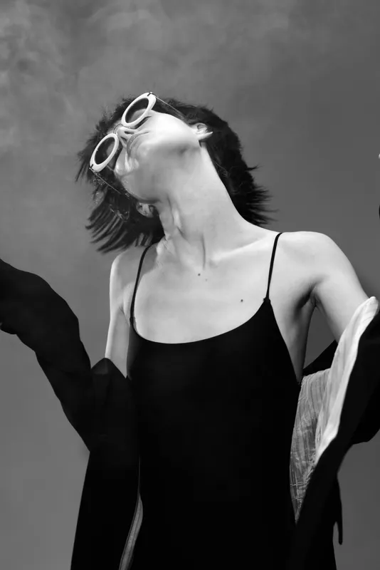 Black and white portrait of a young woman wearing sunglasses in stylish outfit with dynamic pose and expressive mood.