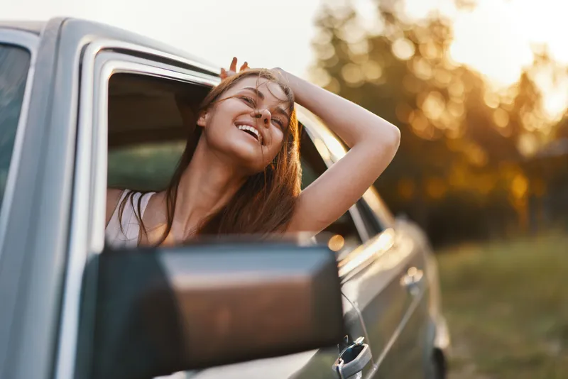 Happy woman enjoying the sunshine from a car window in a lush green setting, embodying freedom and joy in a summer atmosphere.