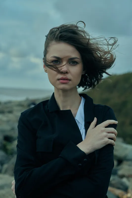 Woman on a windy coast wearing a black shirt, hair swept across her face, moody sky and rugged rocks create a dramatic coastal scene filled with strength and emotion