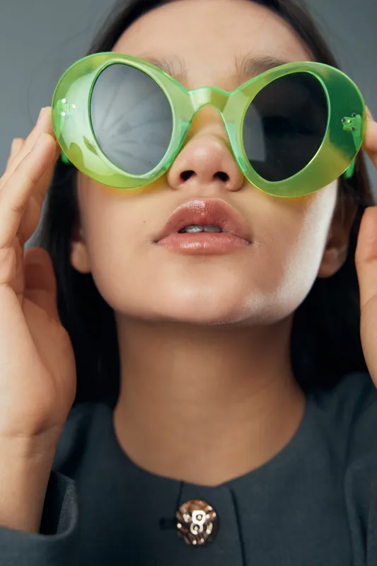 Woman wearing oversized green sunglasses and dark clothing posing indoors with hand adjusting eyewear, fashion portrait with neutral background and glossy lips closeup face style.