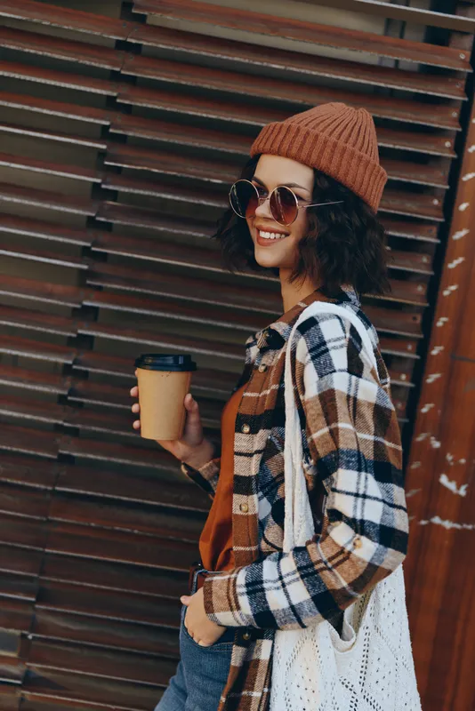 woman smile coffee street autumn beanie sunglasses plaid tote. Urban candid portrait of a young model holding takeaway cup, golden hour glow, authenticity and emotional storytelling for mindful