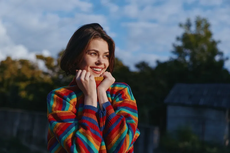 Young woman portrait in colorful striped sweater, smiling and holding collar outdoors in rural setting. Casual outfit, short brown hair, sunlit lifestyle photo for travel and cozy autumn mood.