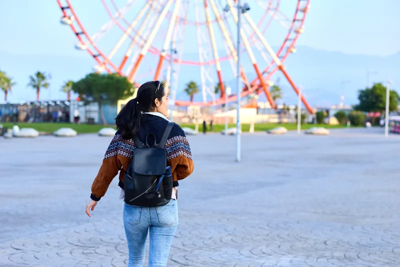 Woman with backpack walking toward a ferris wheel in an outdoor amusement park on a sunny day, exploring park attractions and urban leisure as she wanders along paved paths and observes colorful