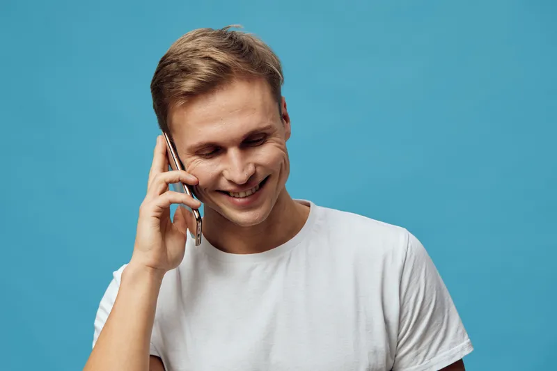 Smiling man talking on mobile phone with cheerful expression, casual white t-shirt, indoor studio shot, vibrant blue background, lifestyle and communication concept