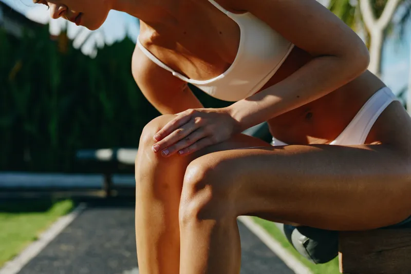 A pregnant woman outdoors squats beside a park path, wearing light athletic wear. The bright natural light highlights maternity strength, lifestyle wellness, and daily fitness in a sunny outdoor