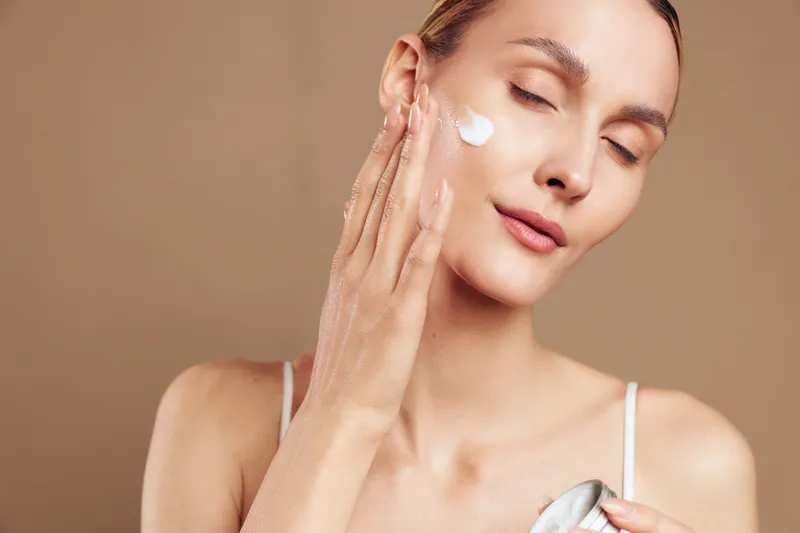 Young woman applying moisturizing cream on face with closed eyes, soft natural light, smooth skin texture, neutral background, beauty skincare routine concept.