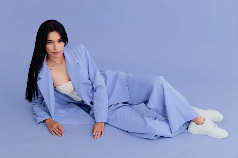young woman in blue suit reclining on floor with white sneakers, long dark hair, calm expression, studio background in soft pastel blue tones