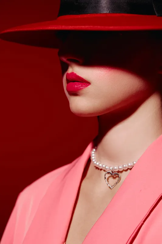 Close-up of stylish woman wearing wide-brimmed hat, bright red lipstick, and pearl necklace with heart pendant. Pink jacket on red background. Creative beauty and fashion concept with shadows.
