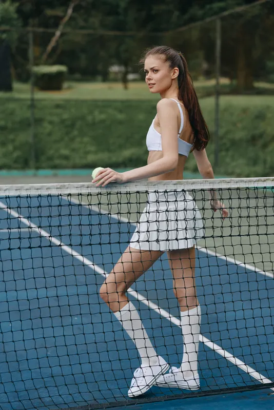 Young athletic woman in sporty outfit posing with tennis ball by the net, showcasing confidence and determination on outdoor court