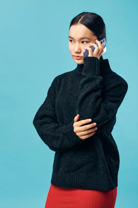 Young woman in black sweater talking on smartphone against blue background. Casual wear, technology use and communication with thoughtful expression and standing posture.