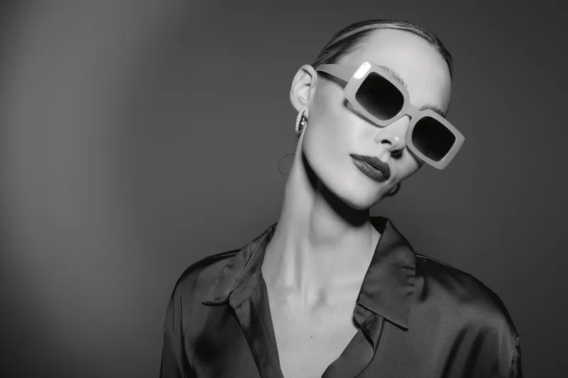 Elegant caucasian woman with pale skin wearing large sunglasses and silk shirt poses against dark background in black and white studio portrait.