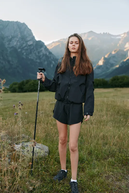 A young woman stands in a grassy field with a trekking pole in hand, set against dramatic mountains. She wears a dark jacket and shorts, looks confident and ready for adventure, capturing a spirit of