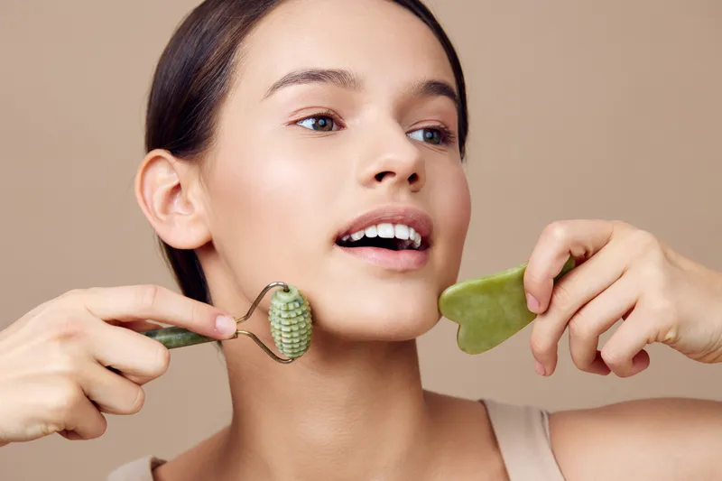 Young woman performing facial massage with jade roller and gua sha tool, promoting relaxation and skincare with a neutral background and natural makeup
