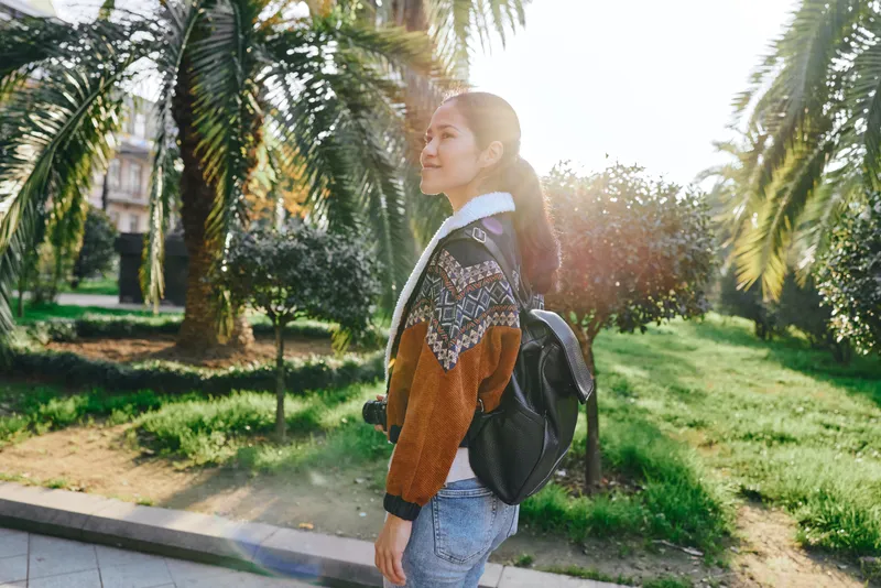 Woman with backpack walking along a sunny park path, casual outfit, palm trees in the background, natural light, candid outdoor moment, travel vibe, serene greenery