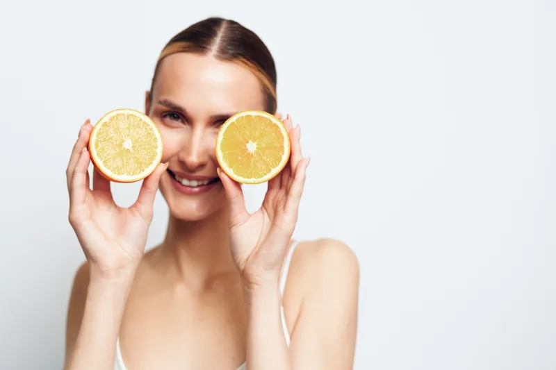 A young woman with long hair holds two halves of a fresh orange in front of her eyes while smiling against a plain white background.