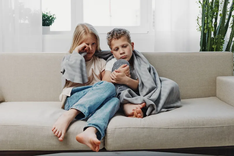 Sad children wrapped in blankets sit closely on beige sofa in bright room, expressing tiredness and comfort. Concept of childhood emotions and family care.