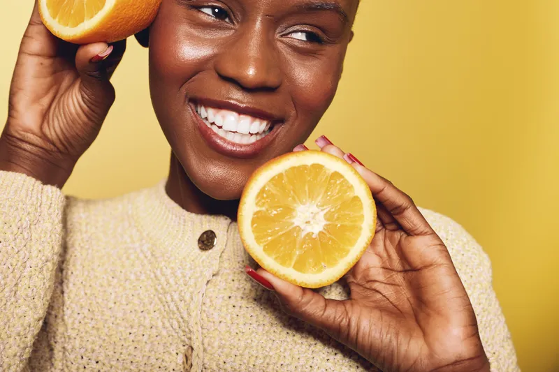 Smiling dark skinned African woman holds fresh orange halves near face on yellow background. Concept of healthy lifestyle, natural vitamins, skincare, and joyful wellness with citrus fruit.