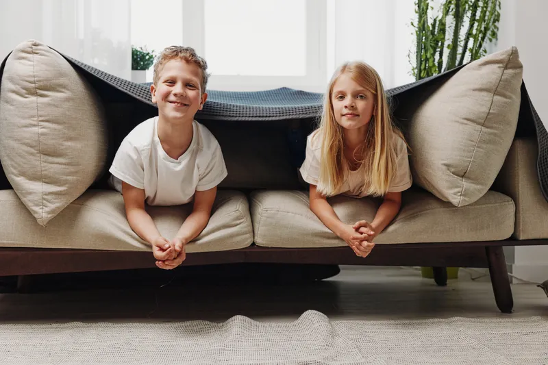 Two smiling children lying under sofa with blanket, enjoying playful moment in bright living room. Concept of childhood fun, family bonding, and indoor activity.