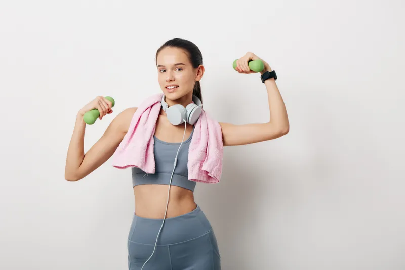 Fitness woman in a cropped top and leggings lifts small dumbbells with a pink towel over shoulders and headphones, posing confidently during a home gym workout.
